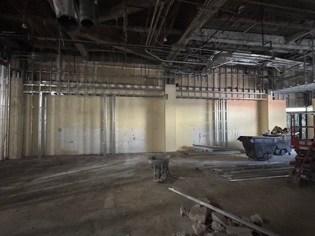 Construction site with unfinished walls, exposed ceiling pipes, and construction materials on the ground.