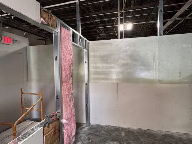 Construction site with partially built interior walls and exposed ceiling, visible pink insulation, and a scaffold in the foreground.