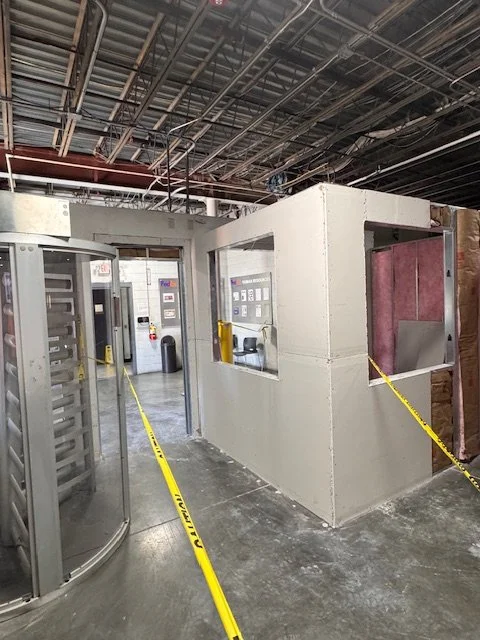 Interior of a storage or processing area with a small white booth, safety yellow caution tape, vending machines, and industrial ceiling.