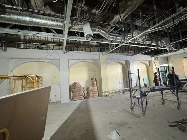 Interior construction site with exposed ceiling ducts, construction materials, scaffolding, and workers.