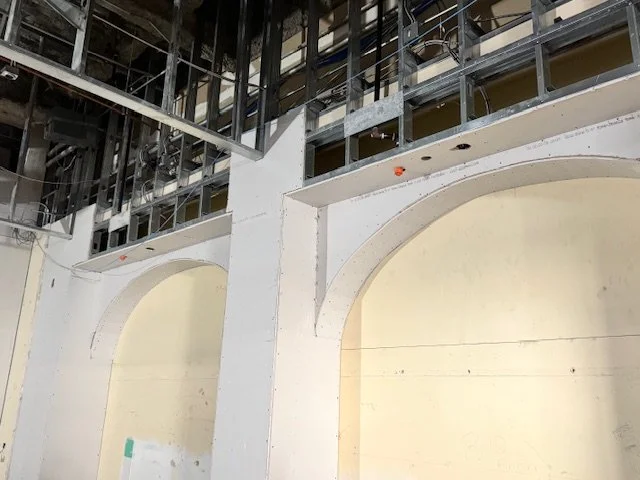 Construction site showing unfinished interior with white walls, large arches, metal framework, and exposed ceiling structure.