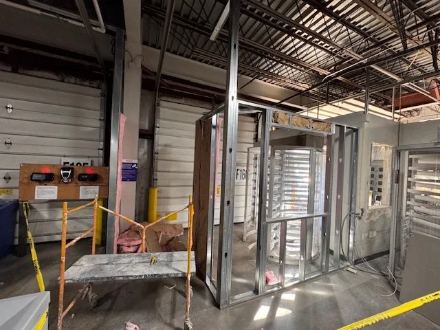 Construction site with metal framing for a new room or wall, surrounded by orange safety barriers and yellow caution tape, inside a warehouse or industrial building.
