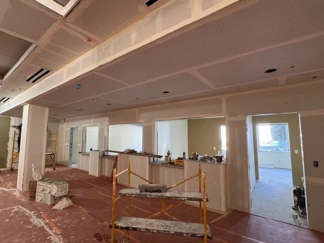 Interior of a building under construction, showing drywall installation, a partially enclosed space with openings, construction tools, and safety barriers.