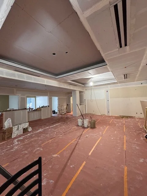 Interior of a building under construction with drywall and ceiling work in progress, construction materials on the floor, and taped markings for flooring or layout.