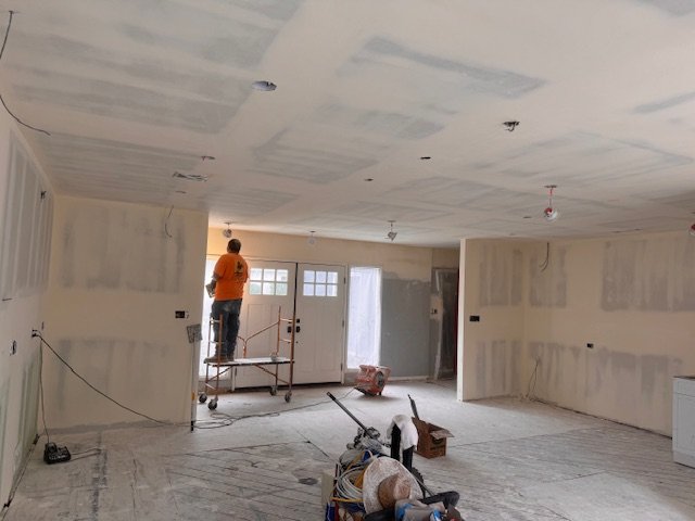 Interior of a house under renovation with drywall process ongoing, worker standing on scaffolding, construction tools and materials on the floor.