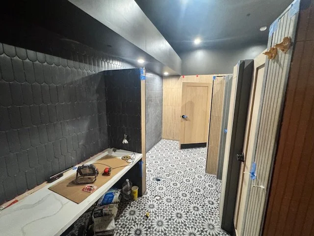 Public restroom with multiple stalls, some doors partially open, black textured wall, patterned tile floor, and construction tools on a counter.