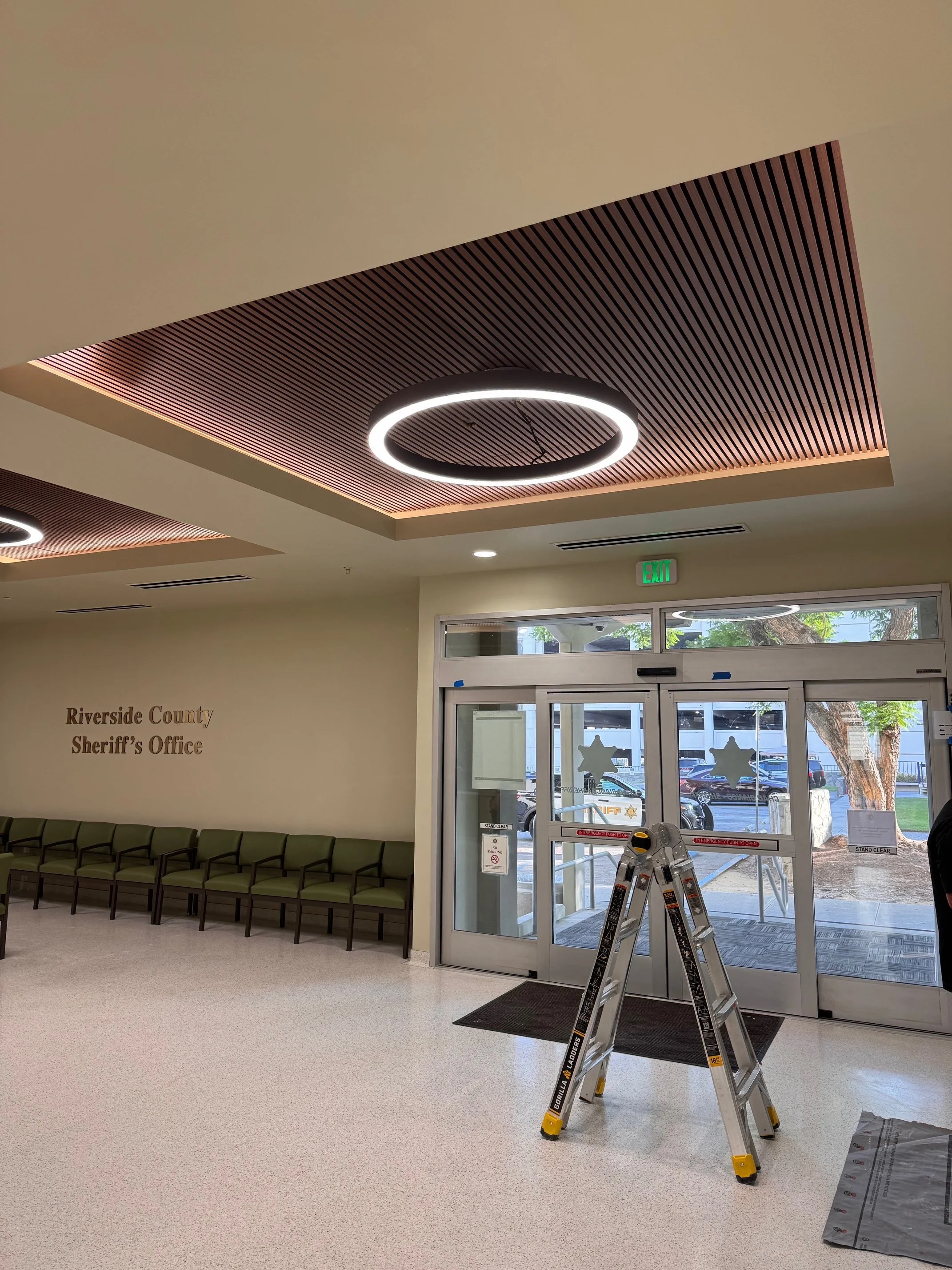 Interior of a public building with a row of green chairs, a decorative ceiling with circular light fixtures, and a glass door entrance. A ladder is set up near the door, and signs and an exit sign are visible.