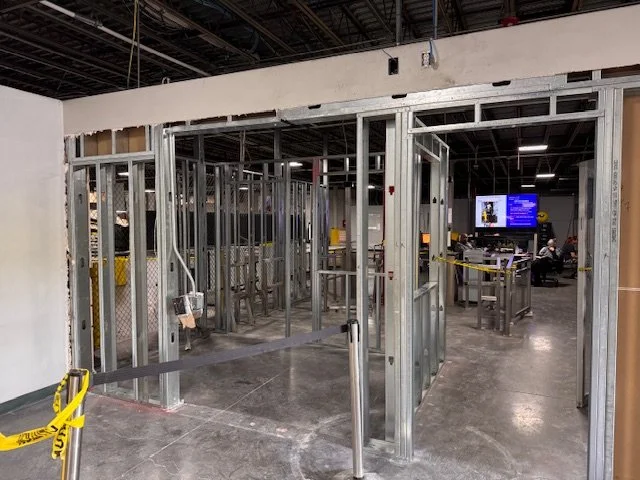 Metal framing for walls or doorways under construction in an indoor space, with yellow caution tape and some workstations or monitors in the background.