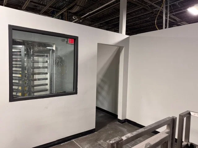 An empty white room with a small window showing an empty, unlit industrial space or kitchen area with metal racks. There is a doorway with no door and a black railing in the foreground.