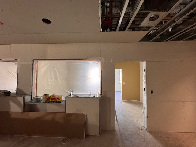 Interior of a building under construction or renovation, with exposed ceiling ducts and pipes, drywall walls, an opening covered with plastic sheeting, and construction materials on a counter.