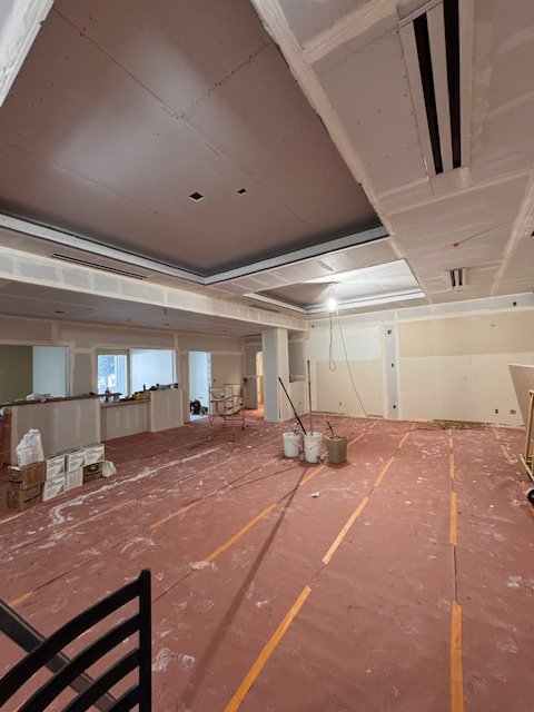 An unfinished indoor space under construction with drywall walls and ceiling, large windows, and a red protective floor covering, with construction tools and materials scattered around.