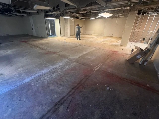 Construction worker standing inside a large, unfinished room with exposed drywall and ceiling, concrete floor, and construction materials around.