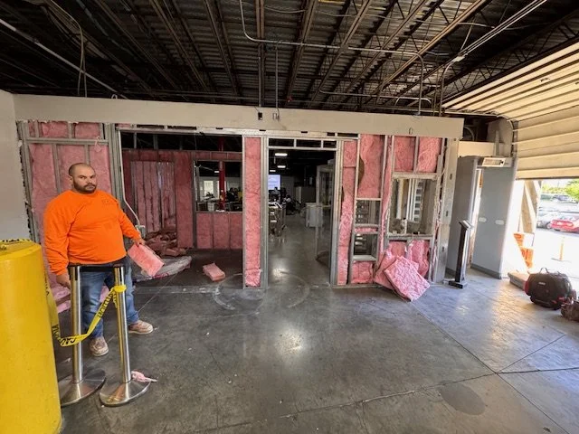 Construction site inside a building with exposed pink insulation and framing, a man in an orange sweatshirt standing near a yellow barrier.