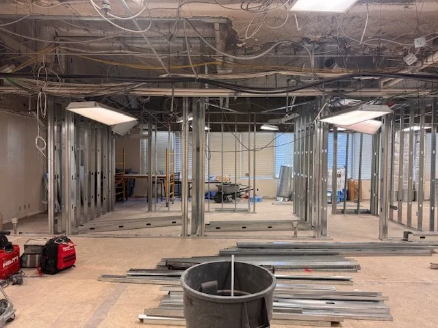 Interior of a building under construction with metal framing, construction tools, and debris, with ductwork and wiring exposed on the ceiling.