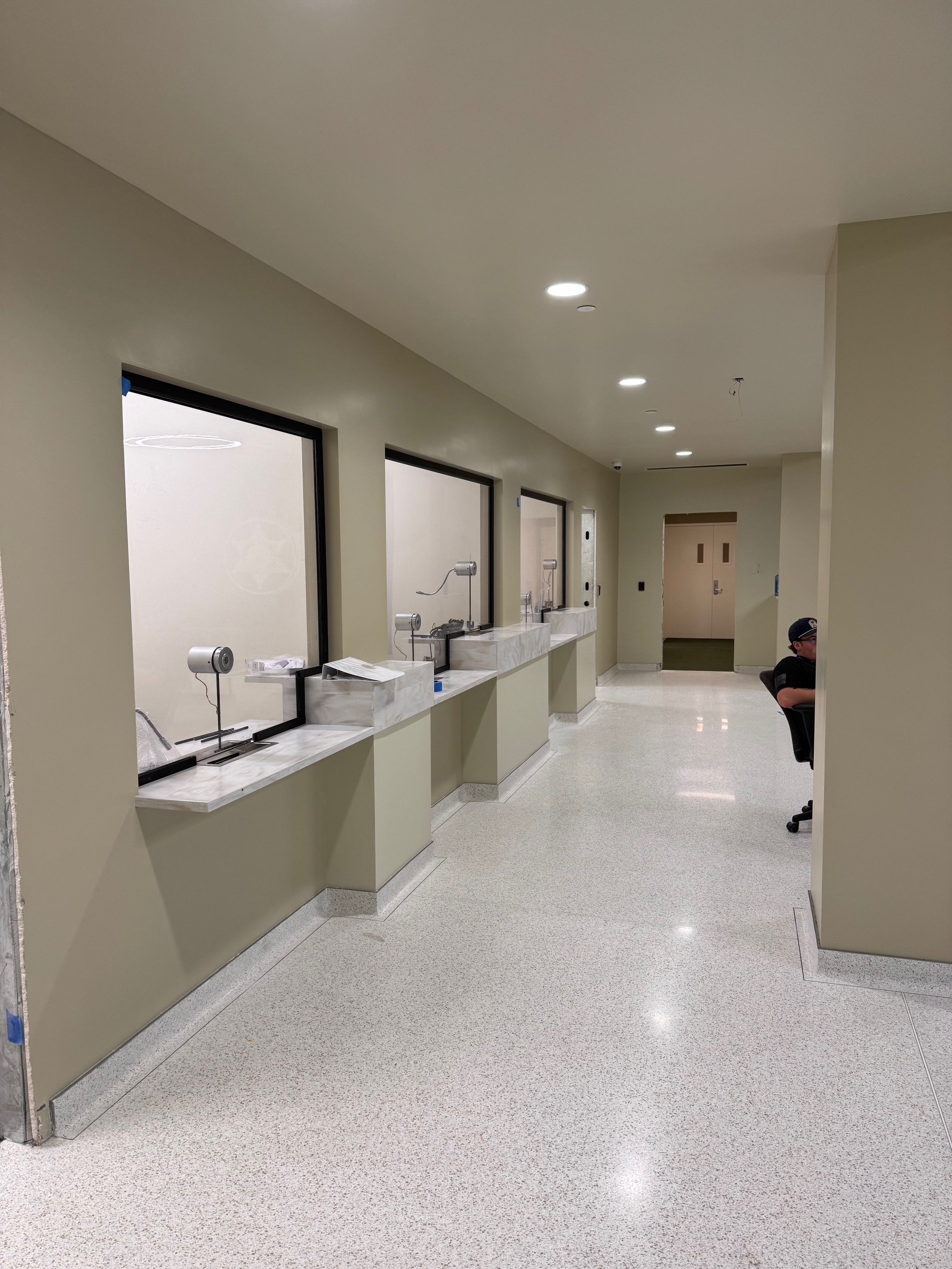 Empty hospital or clinic corridor with windows, sinks, faucets, and a person sitting on a chair wearing a cap, in a clean, well-lit environment.