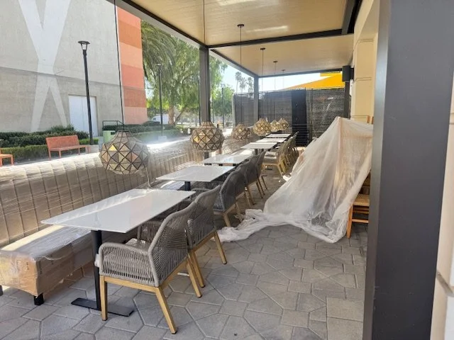 Outside patio with tables and chairs for dining, some furniture covered with plastic, hanging pendant lights, and large windows showing trees and a building.