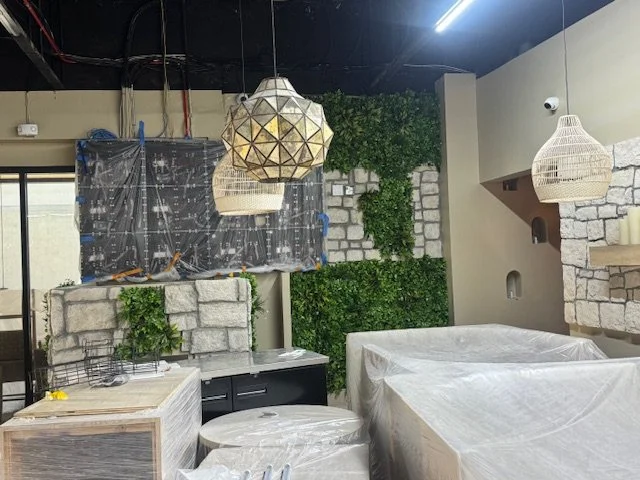 The image shows a room under renovation with hanging light fixtures, a wall with green artificial plants, wrapped furniture, and construction materials.