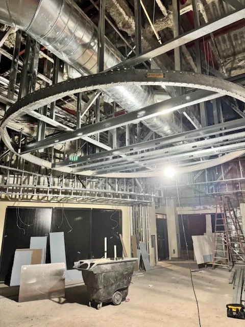 Construction site with exposed metal framing, ductwork, and lights; construction materials and tools on the floor.