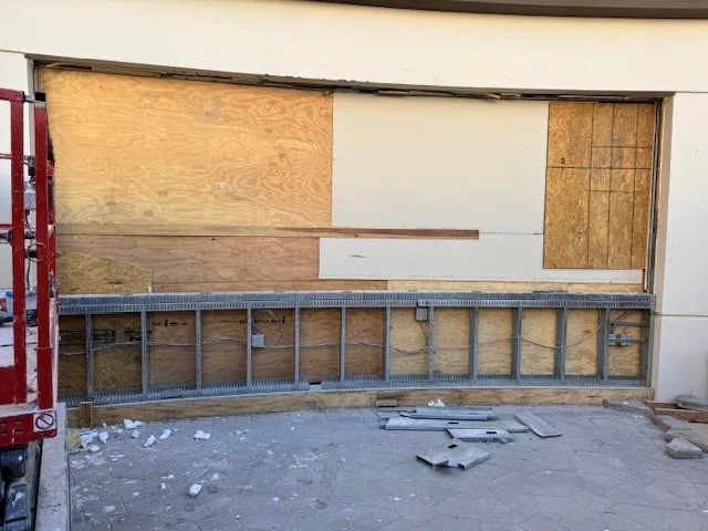 Building under construction with plywood and drywall walls, a metal scaffolding or railing, and construction debris on the ground.