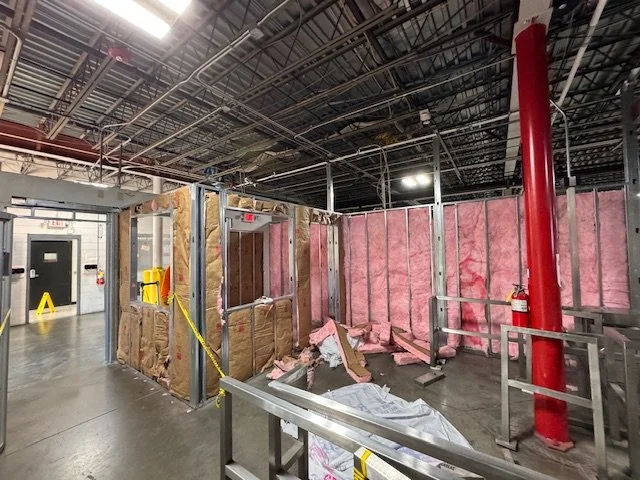 Interior of a building under construction with pink insulation and exposed metal framing, a fire extinguisher on a red pipe, and construction caution tape.