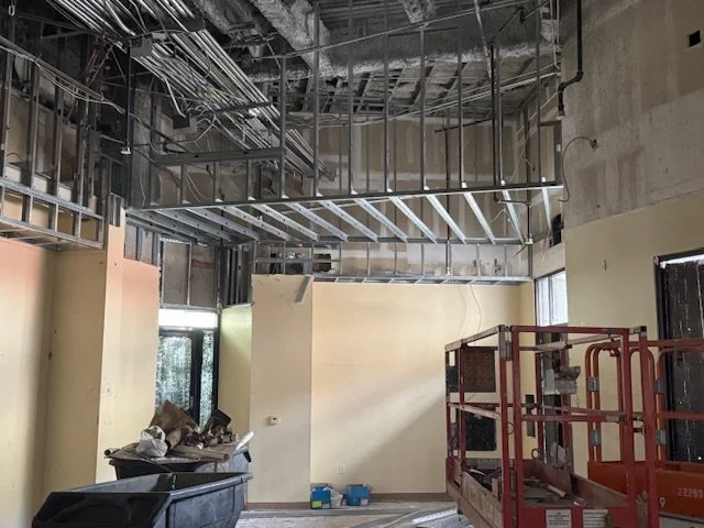 Construction site with exposed ceiling wiring, drywall, and construction equipment including a scaffolding lift and a trash bin.