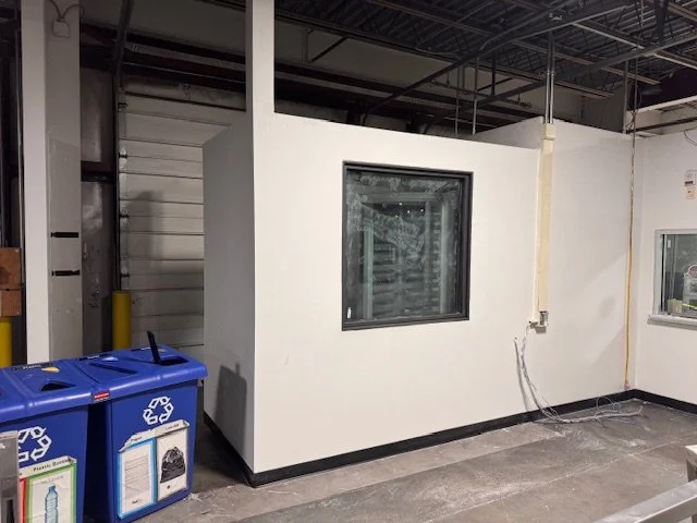 A small enclosed white room or booth with a window inside what appears to be a warehouse or industrial space. To the left, there are blue recycling bins for cans and bottles. Exposed ceiling with metal beams and wiring is visible.