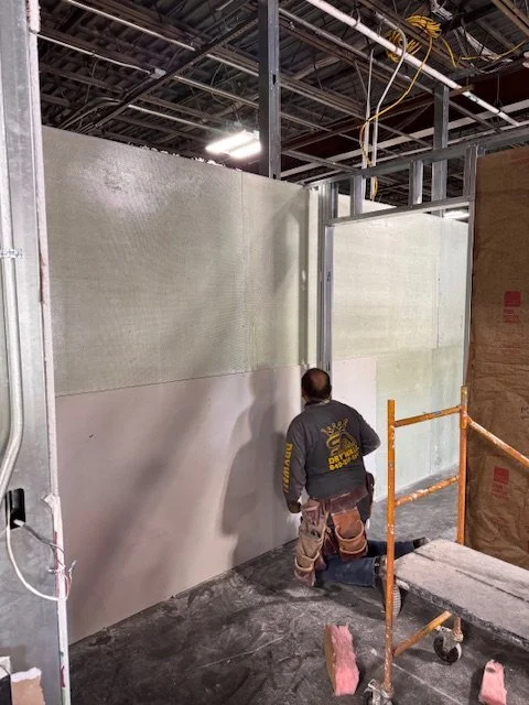 Construction worker installing insulation or drywall panels on a wall in a building under construction, with scaffolding and exposed ceiling pipes.