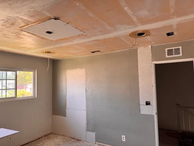 Room under renovation with drywall installation in progress, ceiling with cutouts for lighting, window on left wall, and an opening on the right leading to stairs.