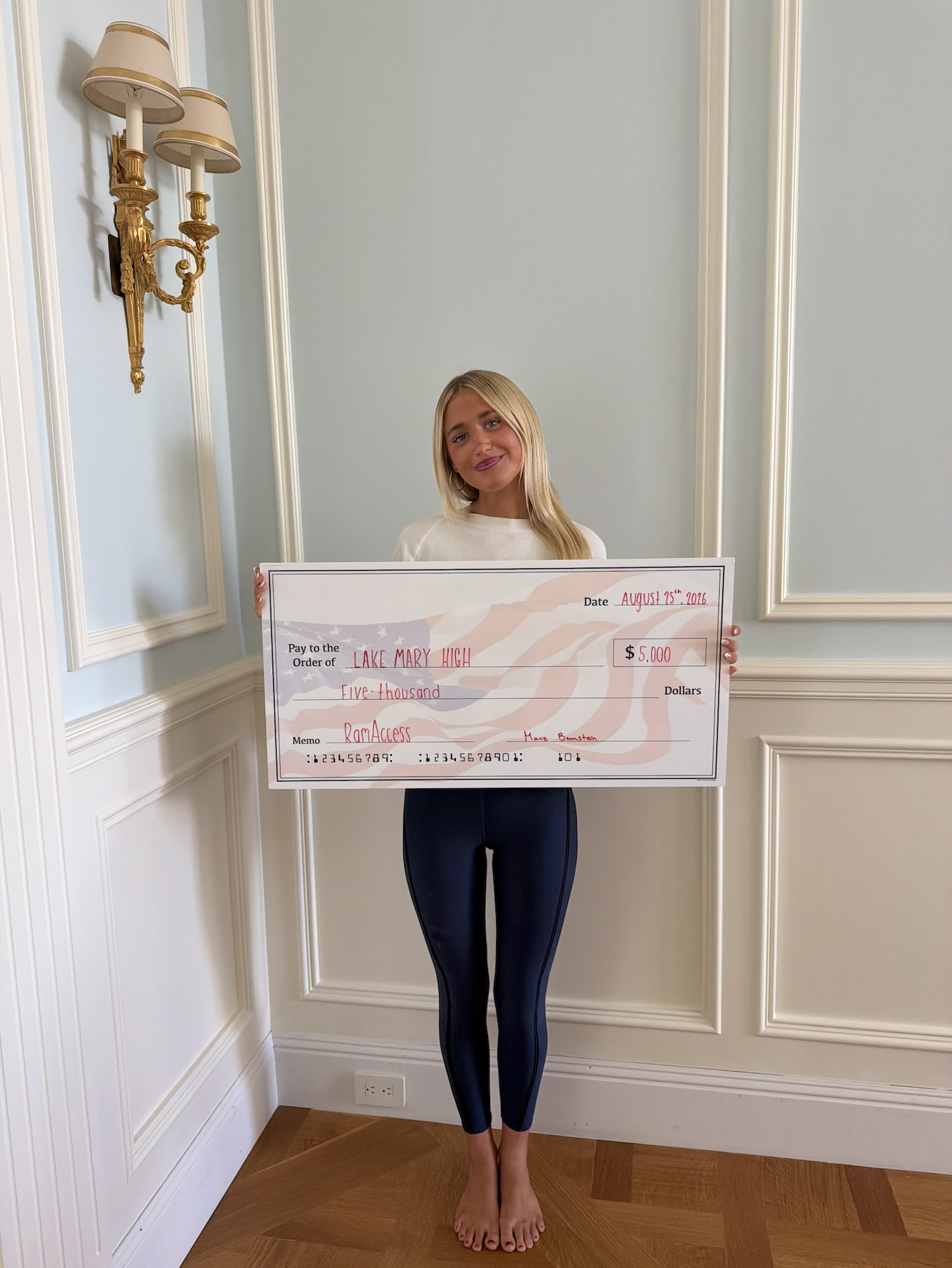A woman standing indoors on a wooden floor, holding a large check, with wall paneling and a gold wall sconce in the background.