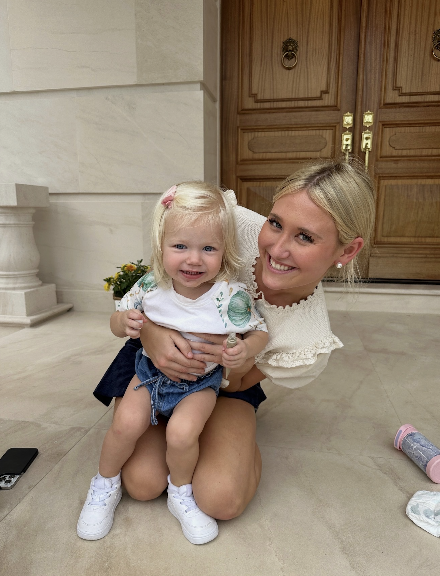 A woman with blonde hair and pearl earrings kneeling on the floor, smiling, holding a young blonde girl with blue eyes, wearing a white shirt with green patterns, denim shorts, and white sneakers, in a room with large wooden doors, a potted plant with yellow flowers, and miscellaneous items on the floor.