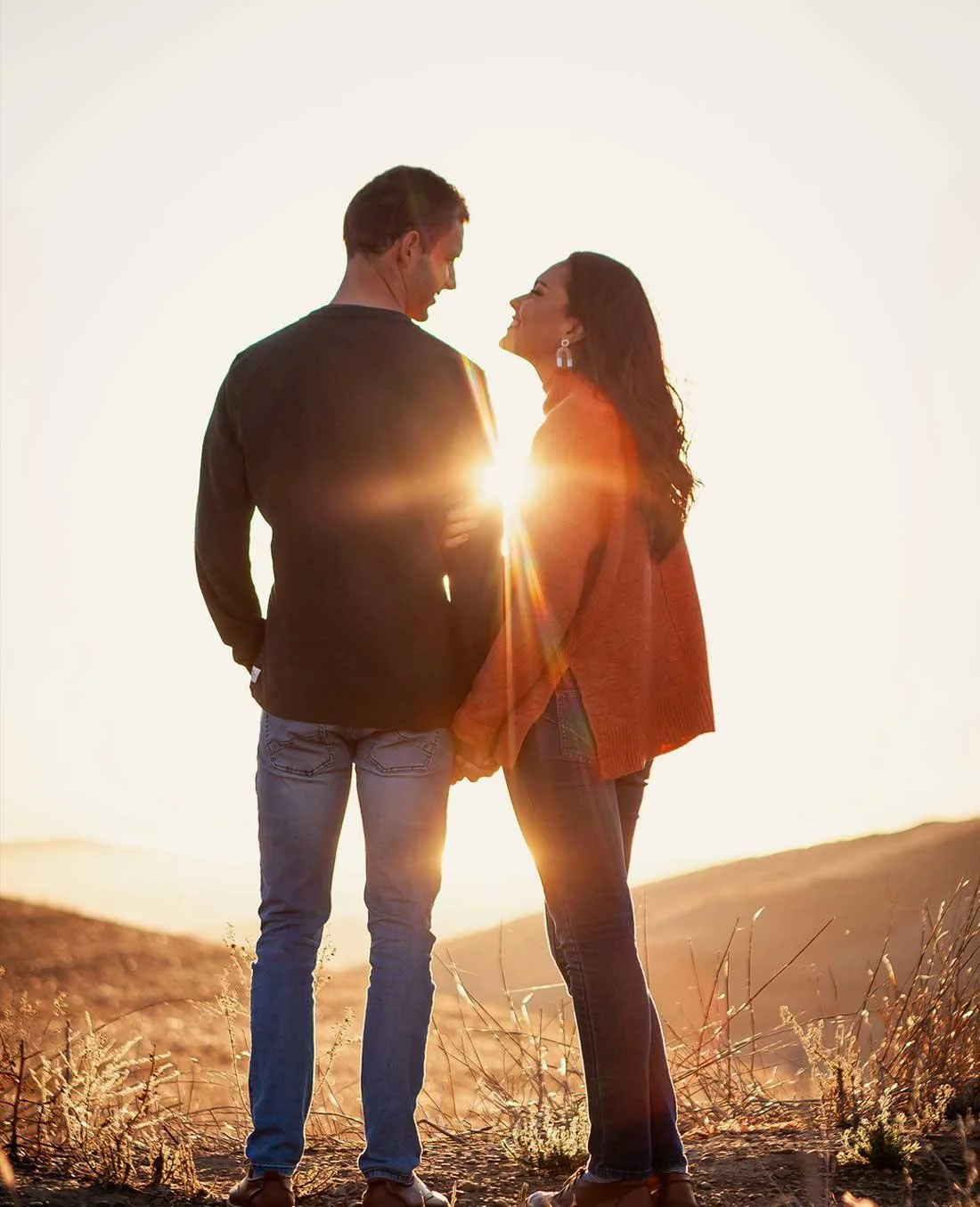 A couple standing hand in hand outdoors at sunset, smiling at each other with the sun shining behind them.