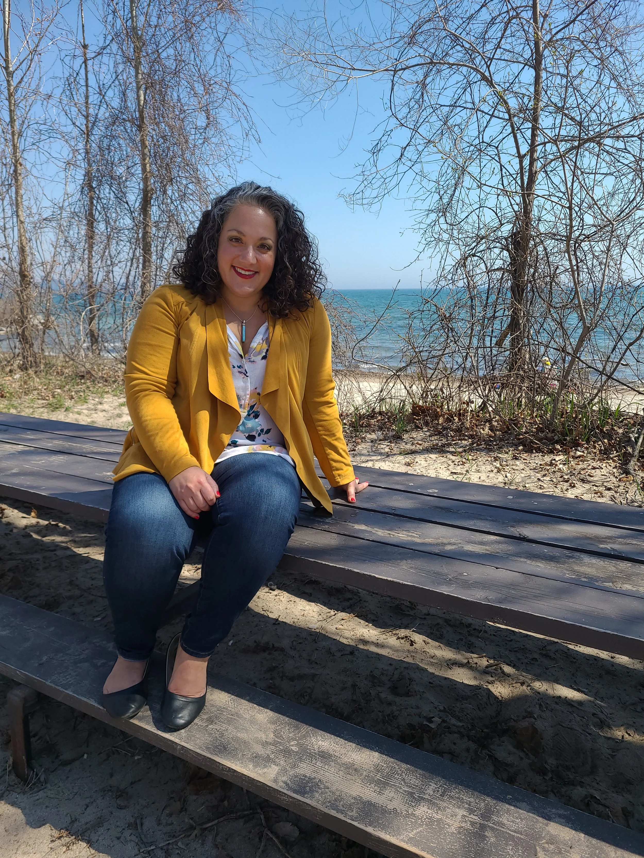 A woman with curly hair wearing a yellow blazer, floral blouse, and dark jeans, sitting on a park bench near a beach, with leafless trees and the ocean in the background on a clear day.