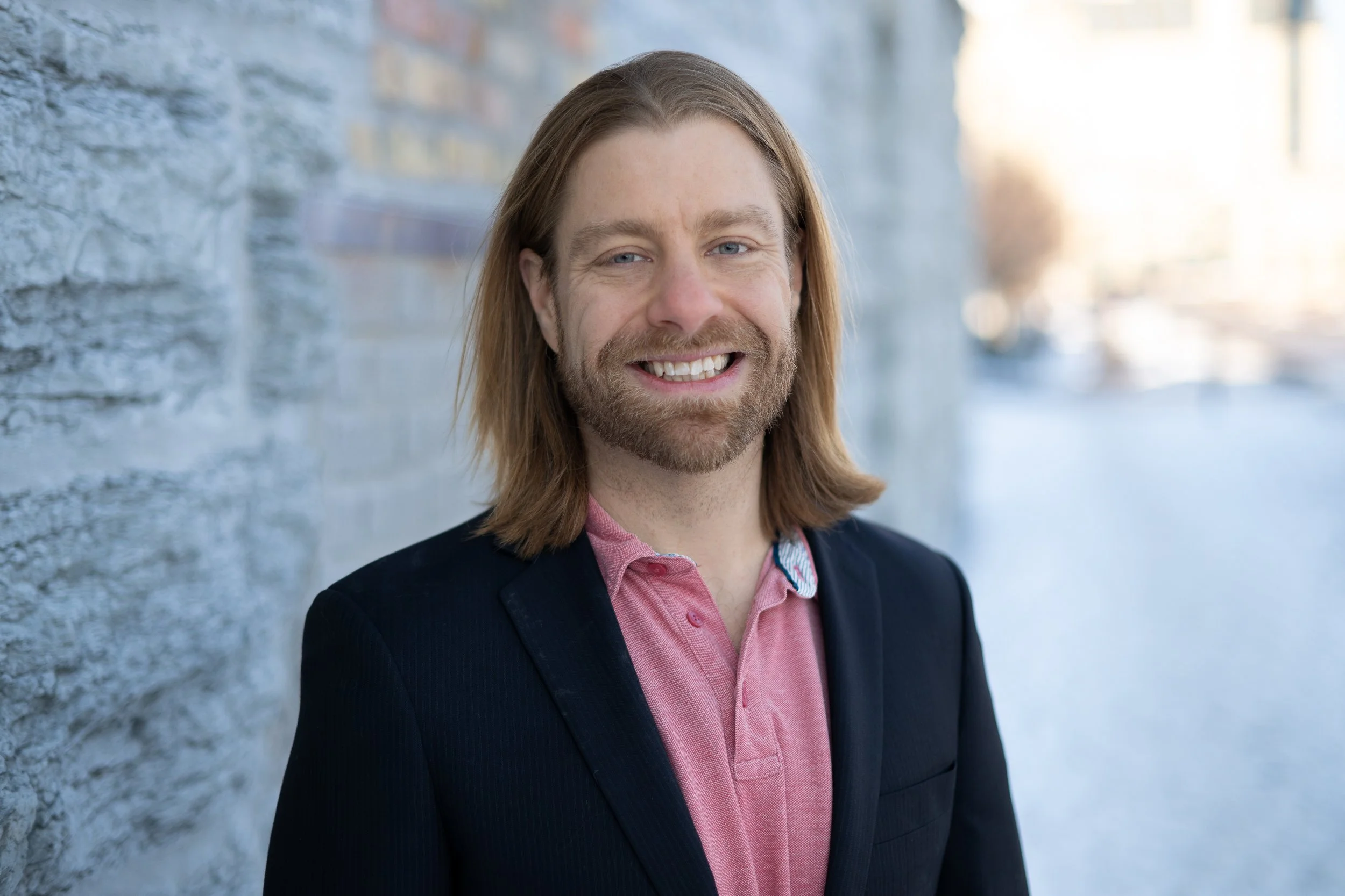 A man with long blonde hair and a beard is smiling, wearing a black blazer over a pink button-up shirt, standing outdoors against a brick wall.