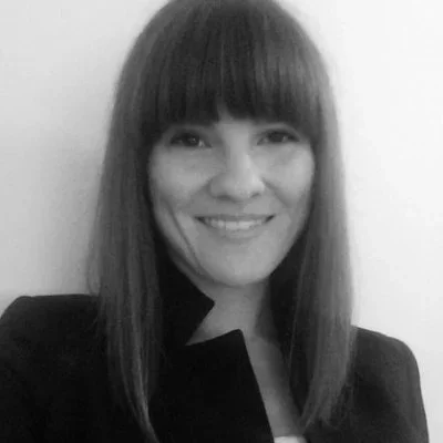 A woman with straight, shoulder-length hair and bangs, smiling, wearing a dark blazer.