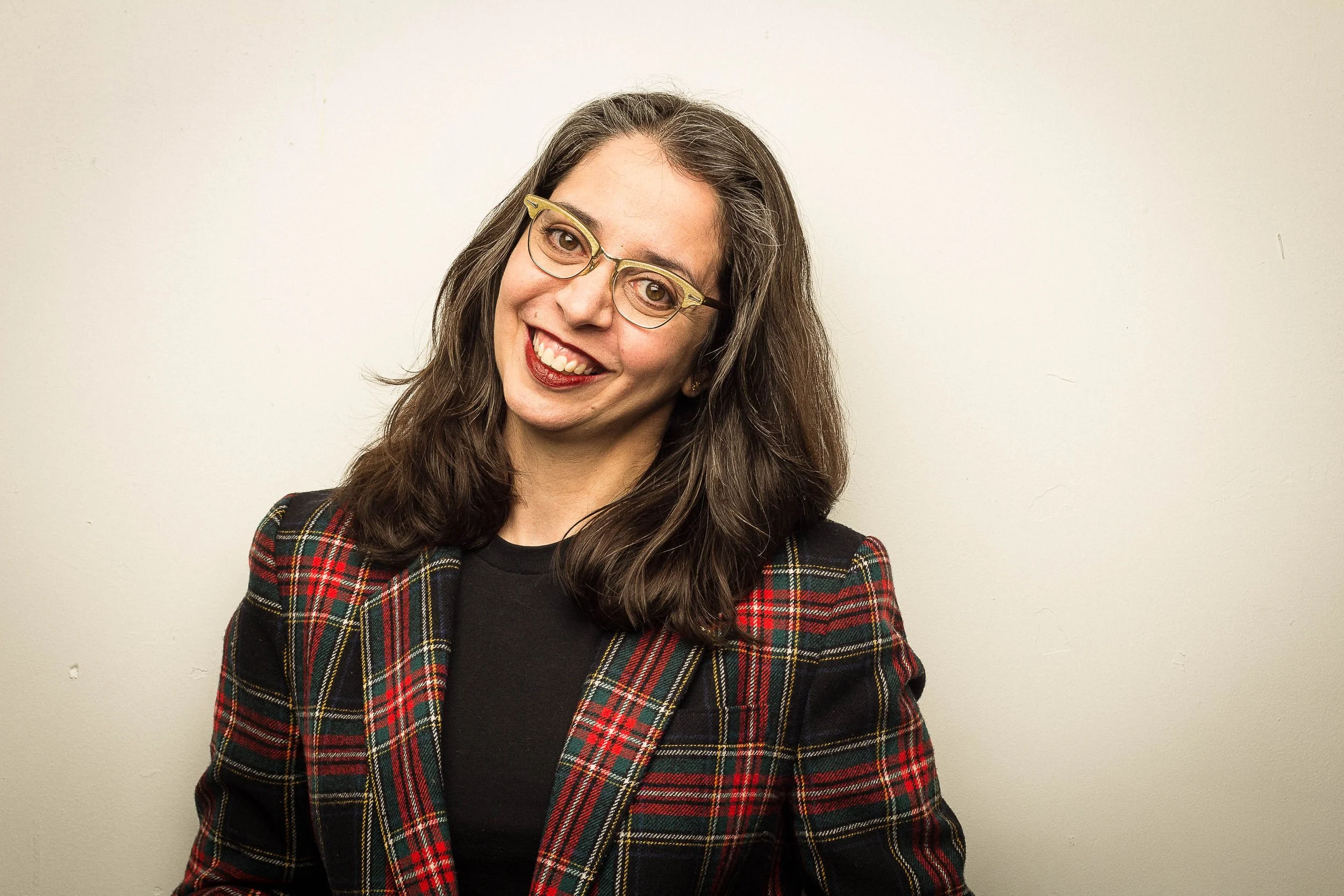 A woman with long brown hair and glasses smiling, wearing a plaid jacket with red, green, and black checkered pattern, standing against a plain white wall.