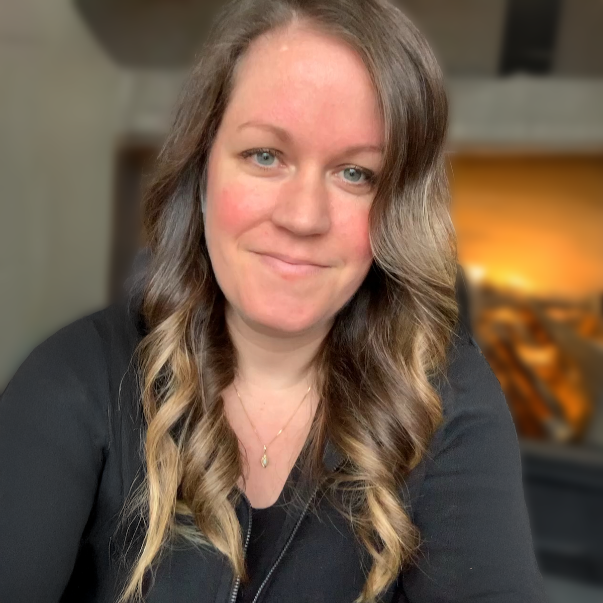 A woman with long wavy hair, wearing a black top and gold necklace, smiling indoors with a blurred background that includes a fireplace and chairs.