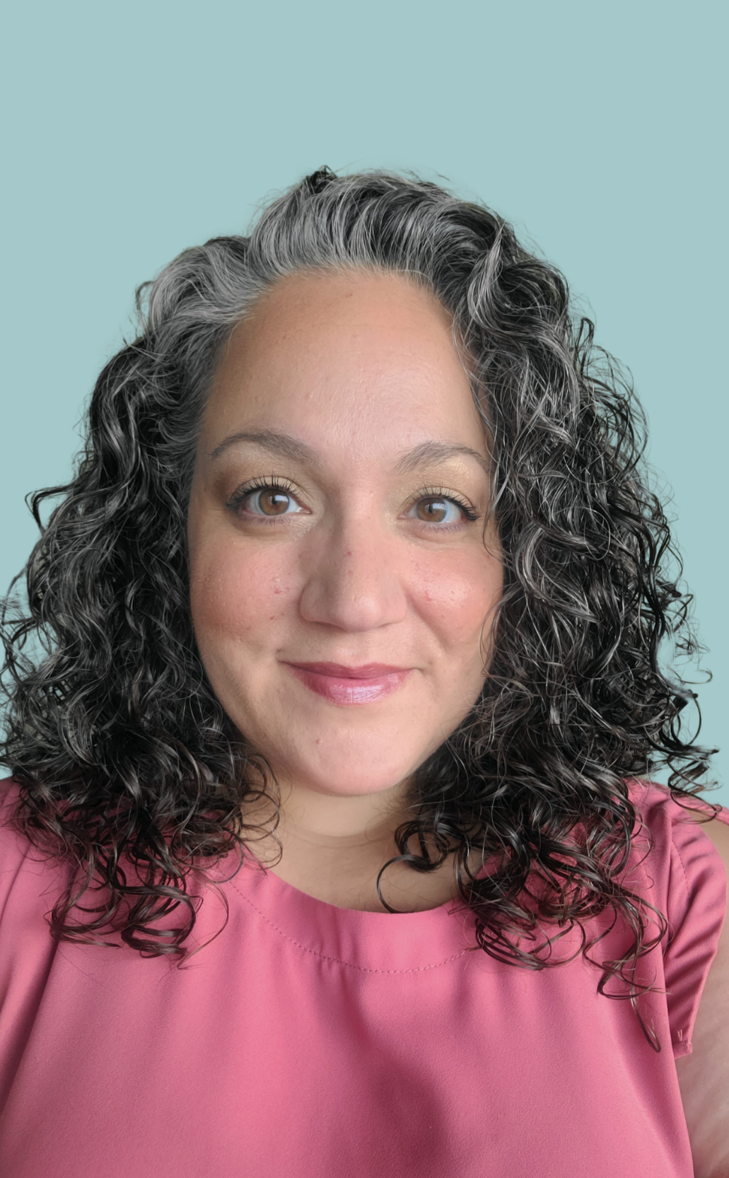 A woman with curly, black and gray hair, wearing a pink top, smiling against a light blue background.