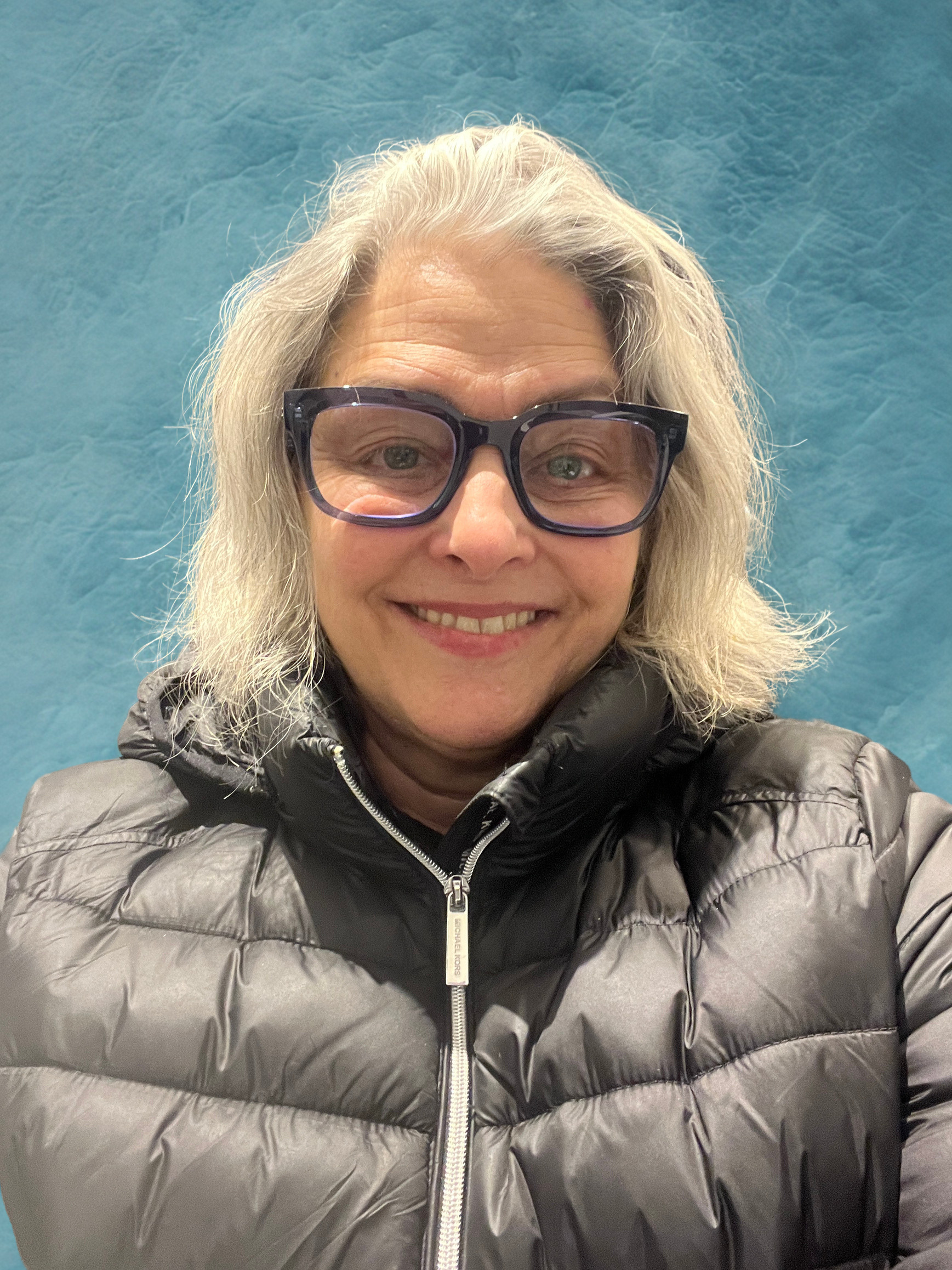 A woman with gray hair and glasses is smiling at the camera. She is dressed in a black puffy jacket with a zipper, and the background is a textured blue wall.