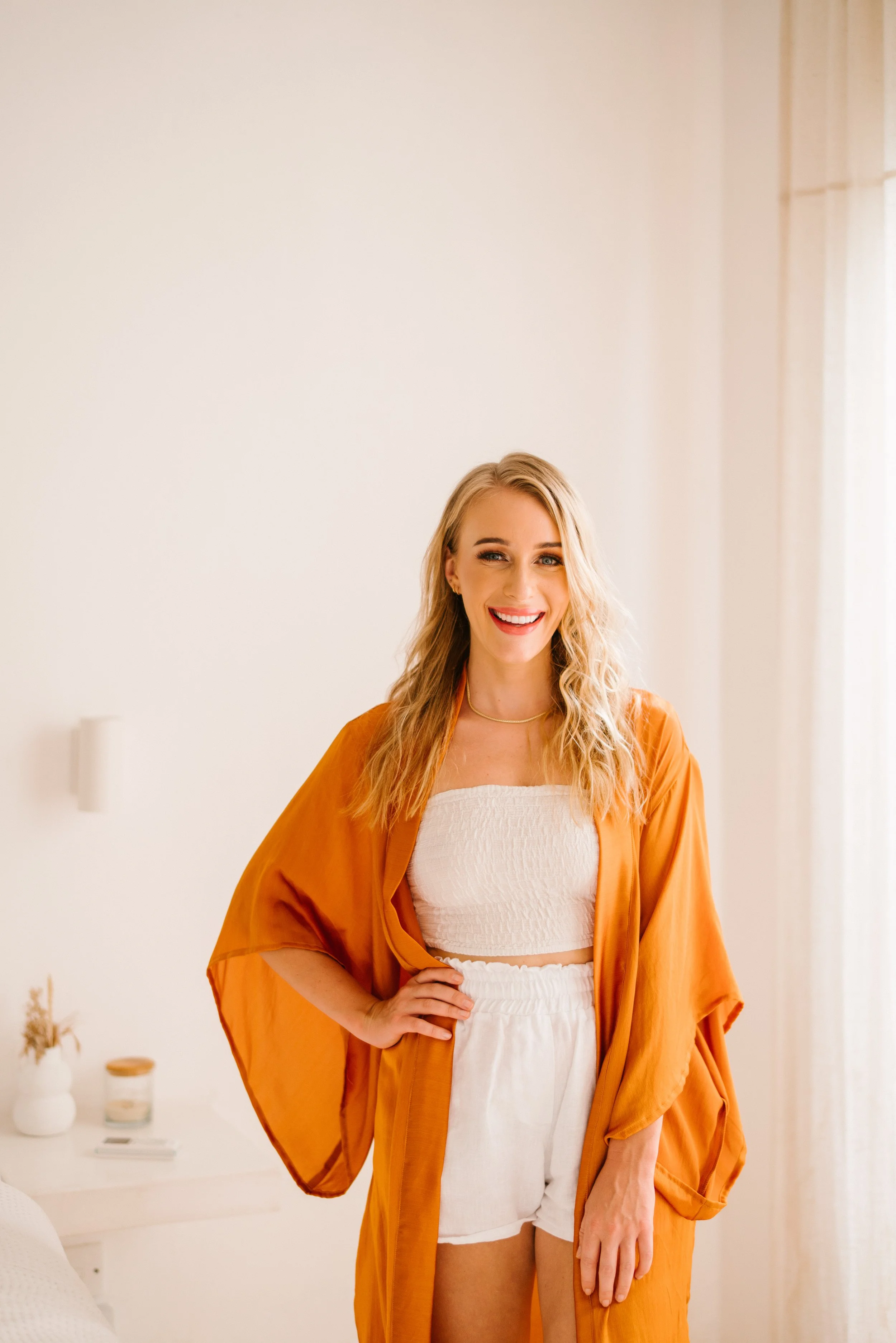 A woman with long blonde hair, smiling and wearing a white strapless top, orange cover-up, and white shorts, standing in a bright, minimalist room.