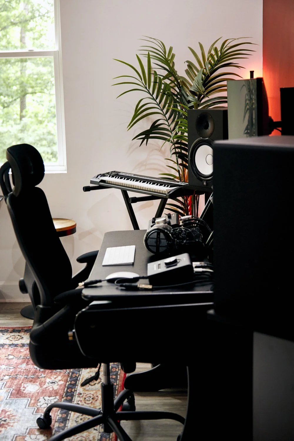 Home recording studio with a black office chair, a computer keyboard and mouse on a desk, audio equipment, a keyboard on a stand, and large studio monitor speakers, with a green plant and natural light coming from a window.