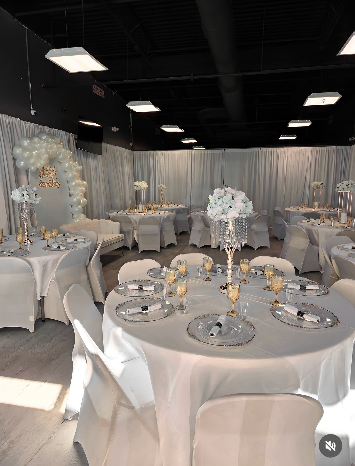 Elegant wedding reception table setup with white tablecloths, gold accents, floral centerpieces, and a balloon decoration arch in the background.