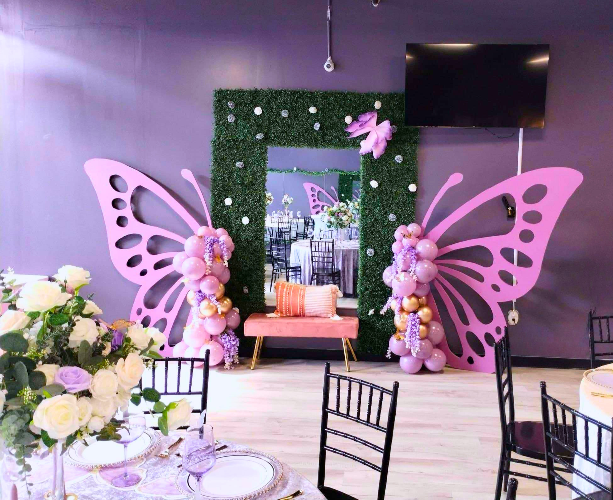 Decorated event space with a green hedge backdrop, pink butterflies, and balloons, featuring pink and white floral arrangements and a small pink couch.