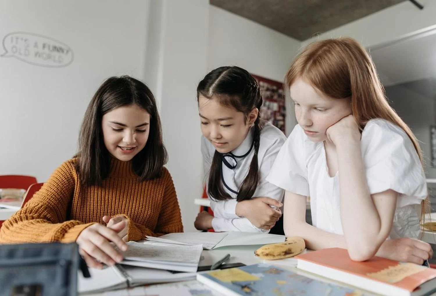 Three young schoolgirls are looking at open notebooks on a table. One girl with dark hair and a brown sweater is pointing at the notebooks, while the other two girls, one with braided hair in a school uniform and the other with red hair in a white shirt, watch intently. A banana and books are also on the table.