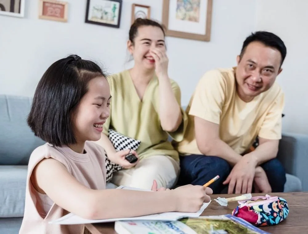 A young girl sitting at a table, laughing and drawing with a pencil, while two adults, a woman and a man, sit on a couch behind her, also smiling and laughing in a casual living room.