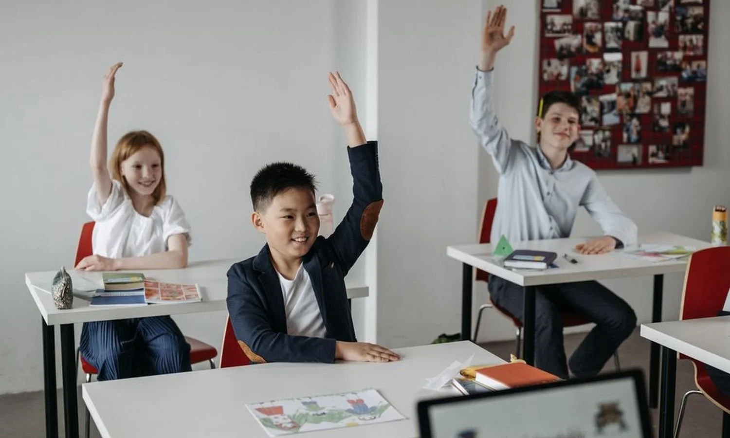 Three children sitting at desks in a classroom, raising their hands.