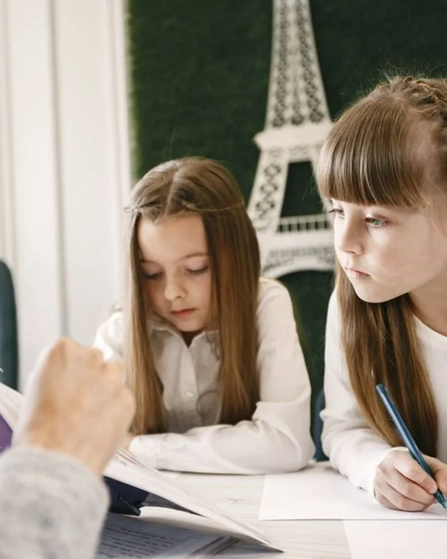 Two young girls with long hair, one with a braid, sitting at a table with papers, writing tools, and a person’s hand pointing at a book, in front of a wall with a partial Eiffel Tower decoration.