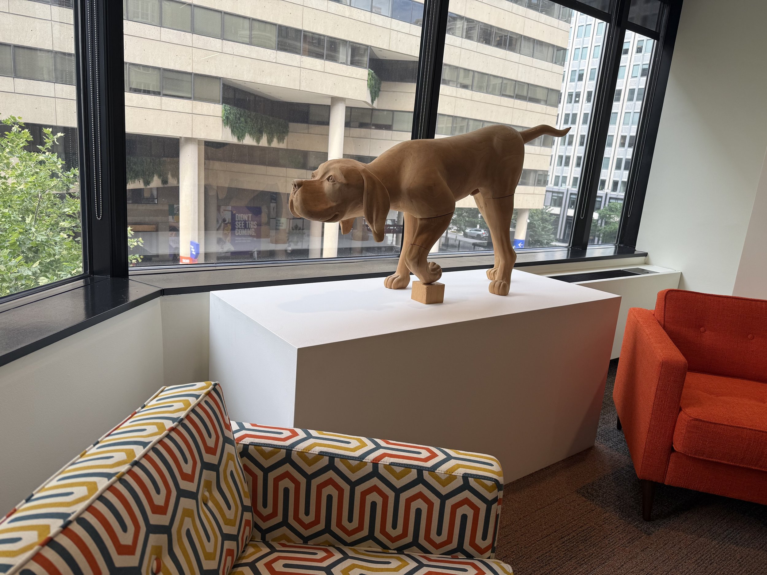 Interior view of an office or lobby with a wooden dog sculpture on a white pedestal near large windows showing city buildings outside. There are colorful patterned and orange chairs in the foreground and a colorful striped cushion.