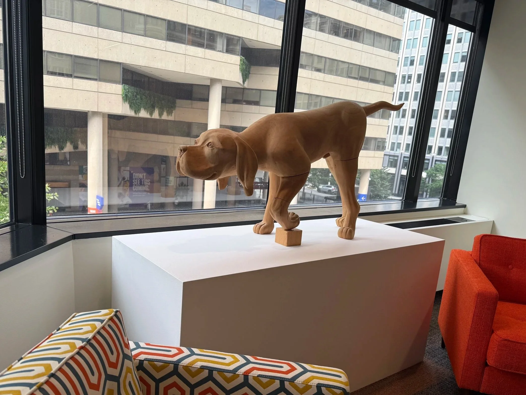 Wooden sculpture of a dog on a white pedestal near a large window in a modern office building.