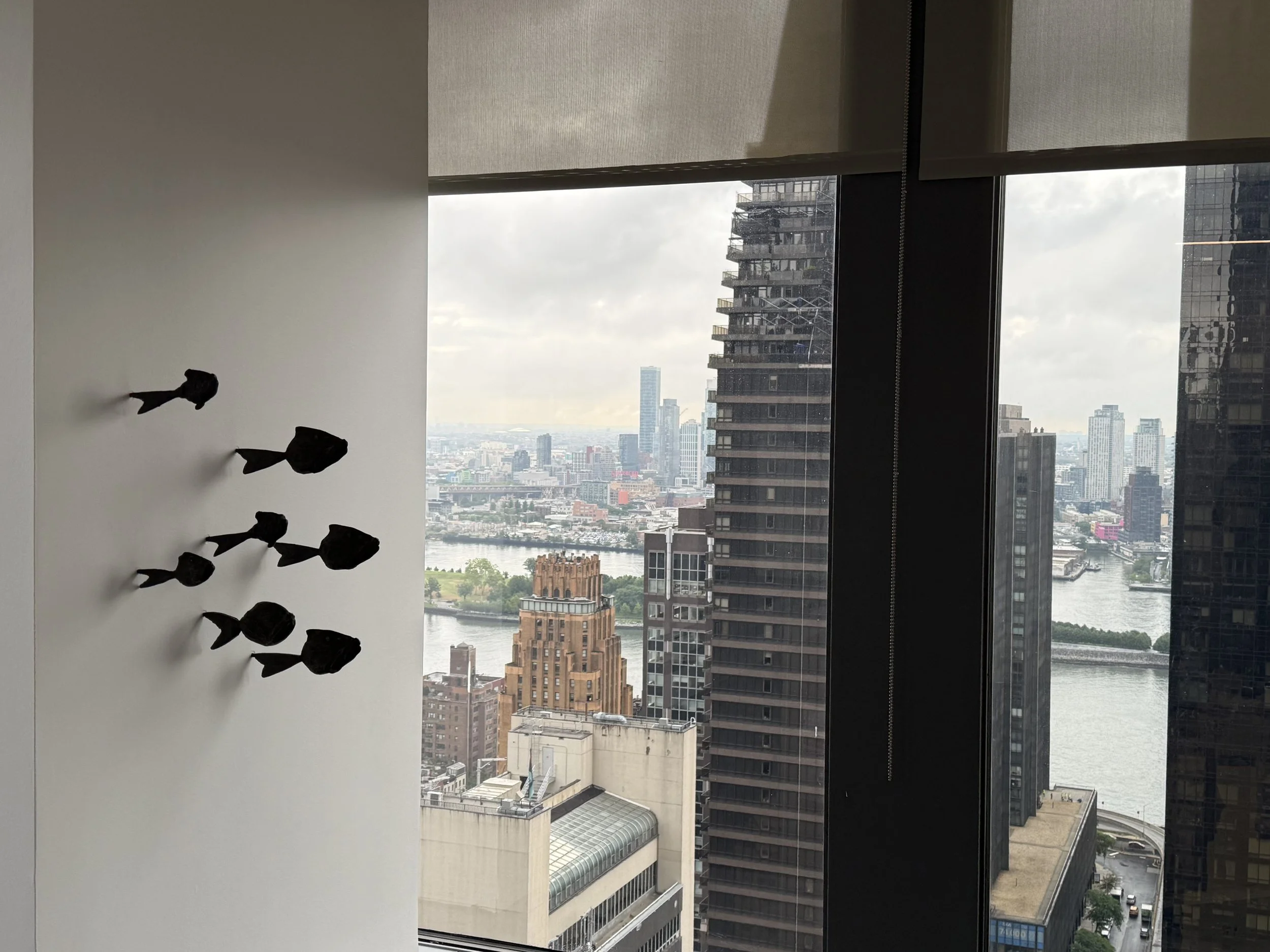 View of city skyline through tall windows, with buildings and a river in the background. A wall with hanging black sculpture in the foreground.