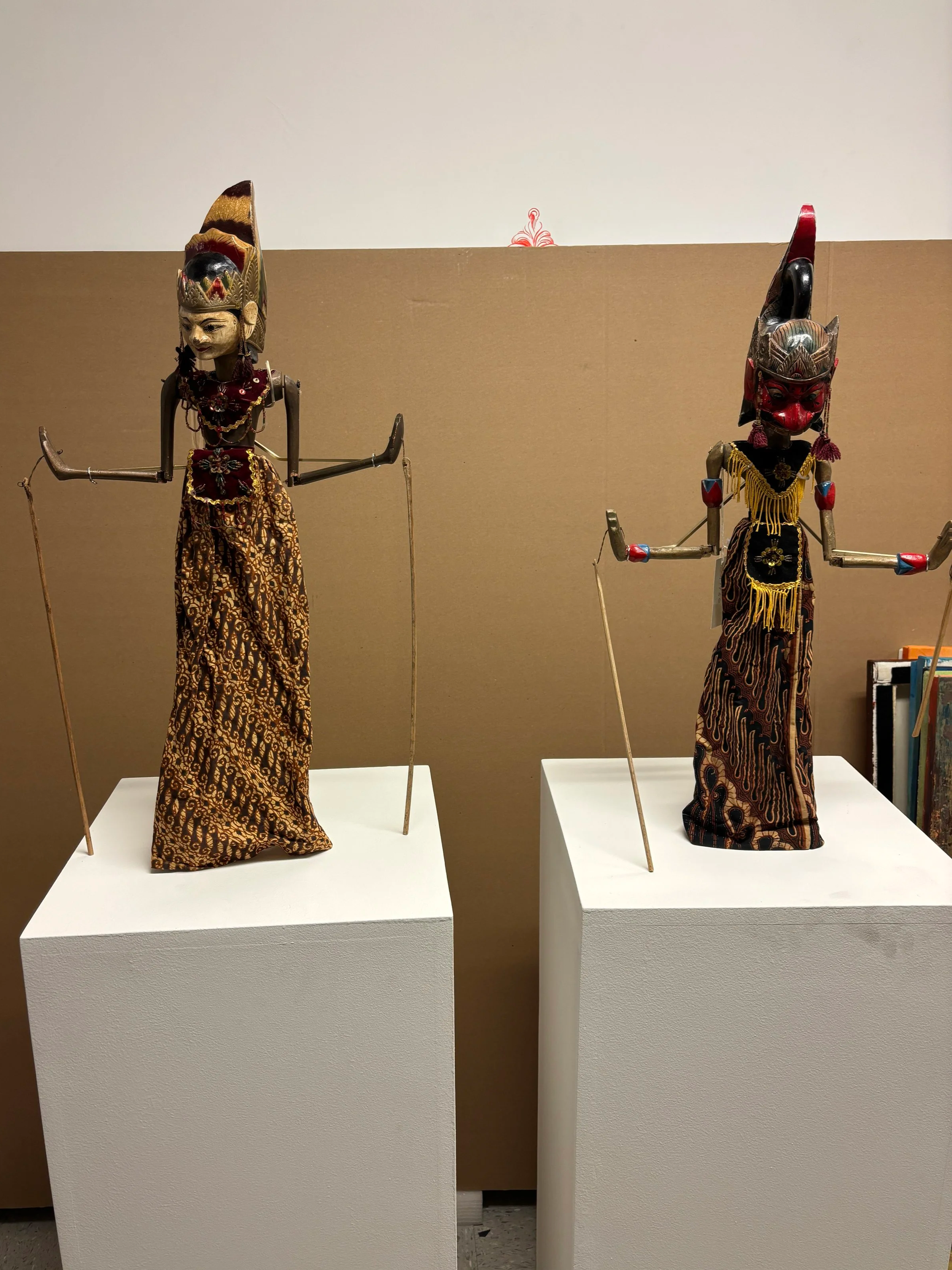 Two traditional shadow puppets displayed on white pedestals against a beige and white background.
